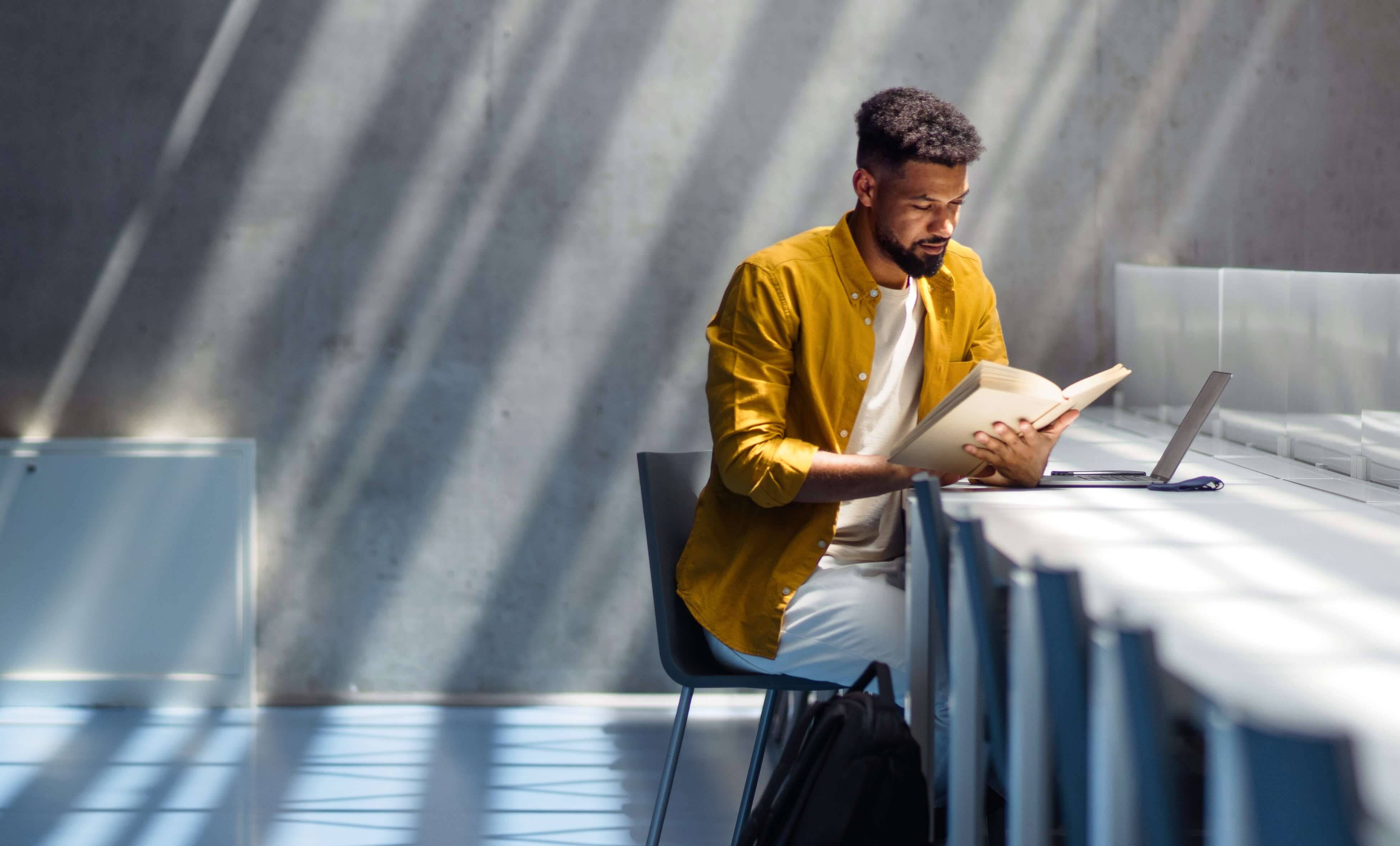 Ein Mann sitzt an einem Tisch, liest ein Buch und hat einen Laptop vor sich. 